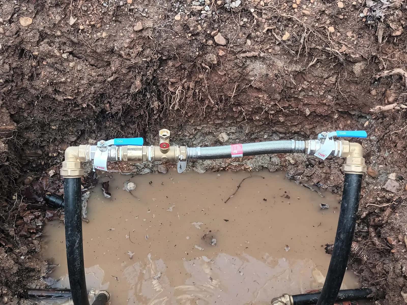 Zanja con tubería nueva, llaves de paso y latiguillo metálico sobre agua embarrada